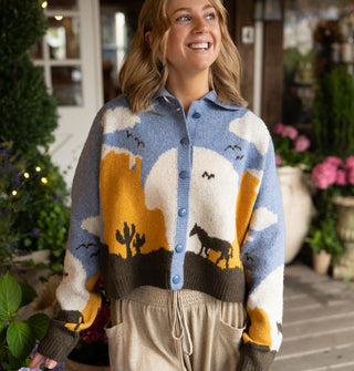 Smiling model wears a button-down collared graphic cardigan with a blue, white, orange, and brown desert landscape scene