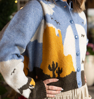 Model wears a button-down collared graphic cardigan with a blue, white, orange, and brown desert landscape scene