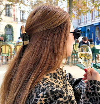 Model wearing a leopard coat and sipping champagne at an outdoor café wears a champagne bottle claw clip in a partially swept-back hairstyle