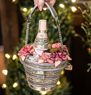 Model's hand holds the strap of a beaded handbag designed to resemble an ice bucket with a bottle of champagne inside flanked by pink flowers