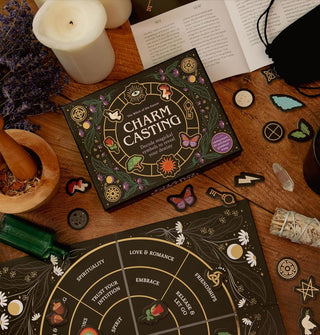 Contents of the Charm Casting kit: board, pieces, guidebook, black velvet drawstring bag, and box on a wooden tabletop staged with candles, sage bundle, mortar and pestle, dried lavender, and a green glass bottle