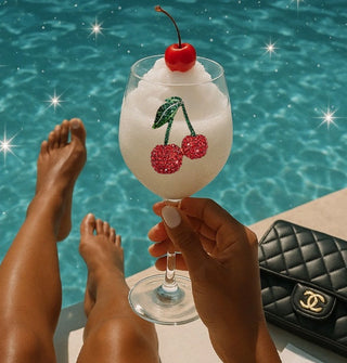 Model holes a rhinestone cherries wine glass filled with a slushy beverage by the side of a pool