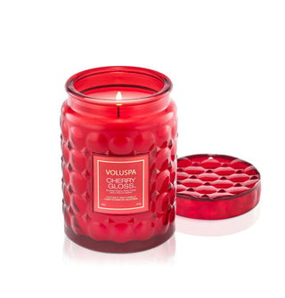 Lit single-wick Cherry Gloss Voluspa candle in embossed red bubble glass vessel with matching lid set to the side