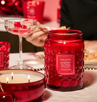 Lit Cherry Gloss Voluspa candles in both bubble glass and red tin vessels staged on a tabletop with cherry-garnished martini glass and a model eating pasta in the background