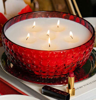 Lit six-wick bowl-style candle in a red bubble glass vessel placed on a round metal tray on top of a book with an opened tube of red lipstick in the foreground