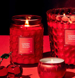Lit single-wick Cherry Gloss Voluspa candle in red bubble glass alongside a larger candle with the same glass and a red tin accent candle in the foreground with glass cherries