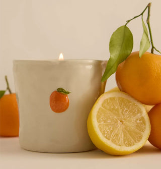 Cream-colored rustic ceramic candle with small sculptural orange fruit embellishment is staged with citrus fruit