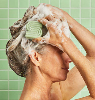 Model lathers hair with an S-shaped ribbed green shampoo bar against a green tiled backdrop