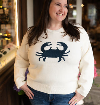 Smiling model wears a cream-colored long-sleeved sweater with central navy blue crab graphic