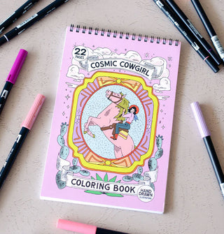 Spiral-bound Cosmic Cowgirl Coloring Book staged with markers features a central illustration of a cowgirl riding a pink bucking bronco