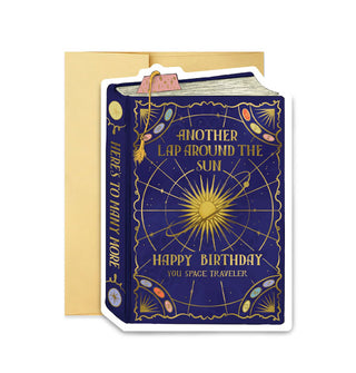 Dark blue greeting card on top of a gold envelope is designed to resemble a book with a gold foil-accented celestial design that says, "Another lap around the sun," and, "Happy birthday you space traveler" on its cover in gold lettering, and, "Here's to many more" on its spine, also in gold lettering