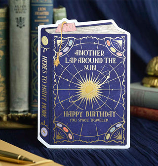 Dark blue greeting card designed to resemble a book with a gold foil-accented celestial design that says, "Another lap around the sun," and, "Happy birthday you space traveler" on its cover in gold lettering, and, "Here's to many more" on its spine, also in gold lettering