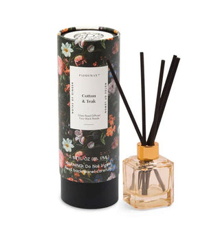 Peach-colored square glass diffuser bottle with several black reeds emerging from the top is placed next to a black floral-patterned tube labeled, "Cotton & Teak"