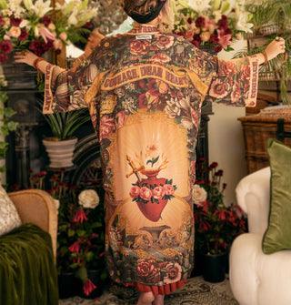 Model with arms outstretched wears a long robe with sacred heart design surrounded by lush florals and the words, "Courage, dear heart" in a banner near the shoulders and on the cuffs