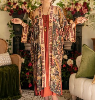Model with arms outstretched wears a long robe with lush floral print and the words, "Courage, dear heart" along its border and cuffs