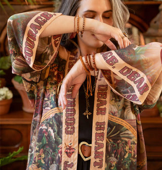 Model with arms upraised wears an intricately patterned open-front tunic with a border that says, "Courage, dear heart" in large lettering