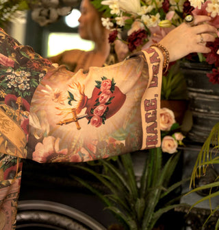 Model with arm outstretched wears a tunic with wide sleeve featuring a sacred heart design and cuff that says, "Courage, dear heart"