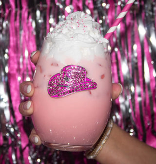 Model's hand holds a stemless wine glass with a pink rhinestone-encrusted cowboy hat decal and filled with a pink beverage topped with whipped cream against a backdrop of pink and silver tinsel