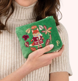 Model holds a small green velvet pouch that features embroidery depicting a fox wearing coat, boots, scarf, and mittens encircled by fall leaves