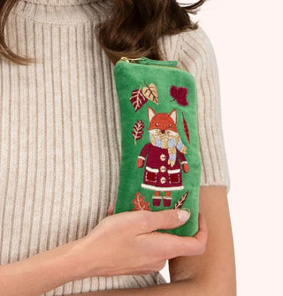 Model in beige ribbed sweater holds a rectangular green velvet eyeglass case with gold zipper and front embroidery of a fox wearing a coat, scarf, mittens, and boots, encircled by autumn leaves