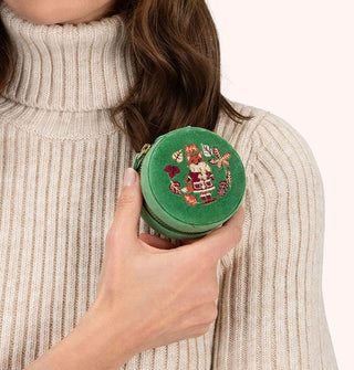 Model in beige ribbed turtleneck sweater holds a round green velvet case with gold zipper and embroidery on its lid that features a fox wearing a coat, scarf, boots, and mittens, encircled by fall leaves