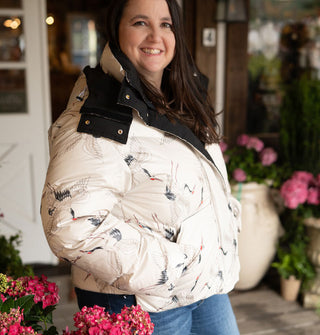 Smiling model stands with one hand in the pocket of a white puffer jacket with black lining and details and all-over delicate print of cranes in flight