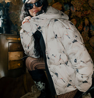 Crouching model in dark sunglasses wears a puffer-style jacket with hood featuring an all-over print of cranes in flight
