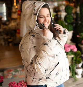 Smiling model holds either side of the collar of a white puffer jacket with hood up that features a delicate all-over pattern of cranes in flight