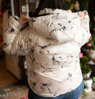 Model facing away from the camera wears a hooded white puffer jacket with all-over print of cranes in flight