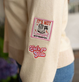 Closeup of two patches on the sleeve of a cream-colored sweater: one says, "It's not easy being a cowgirl but if the boots fit" and the other says, "Let's go girls"