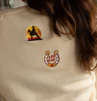 Two embroidered designs on the back of a cream-colored sweater feature a cowboy riding a horse silhouette against the sunset and the other says, "Yee Haw" inside a horseshoe