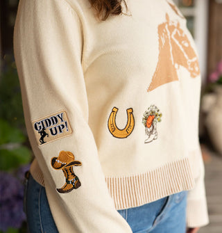 Patches on the front and sleeve of a cream-colored horse head sweater feature Western-themed designs like cowboy boots, horseshoe, and "Giddy Up!" bucking horse