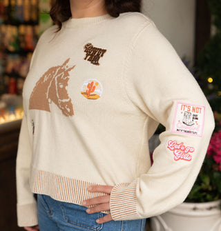 Model with hand on hip wears a cream-colored sweater with central woven horse head design and scattered cowgirl-themed patches