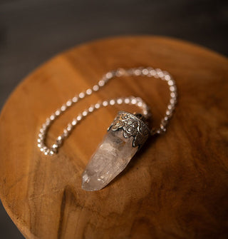 A large clear crystal point pendant in a decorative sterling silver setting with chain attached rests on a round wooden tabletop
