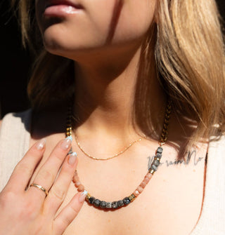 Model lightly touches the necklace she is wearing that is strung with faceted charcoal-colored larvikite stones and peach moonstones with brass separators on a chunky brass curb chain