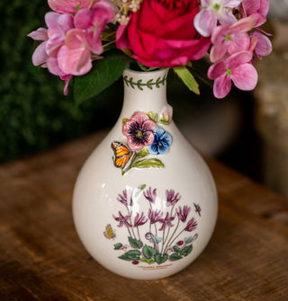 White teardrop-shaped ceramic vase features illustrated design of purple cyclamen below a three-dimensional pansy and butterfly accent and green vine border around the top opening, out of which a bouquet of pink flowers emerges