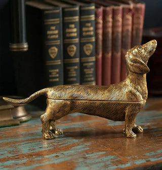 Gold-toned decorative box shaped like a dachshund on a distressed wooden tabletop with books in the background