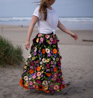 Model wears a black skirt embroidered with colorful, all-over three-dimensional flowers on a windy beach