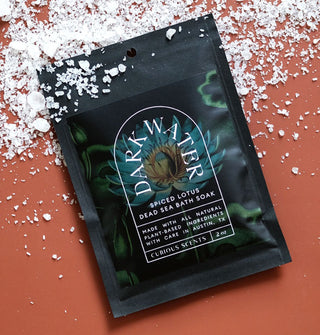 Black packet of Dark Water Spiced Lotus Dead Sea Bath Soak with dark blue lotus and lily pad motif and white lettering rests on a terracotta-colored surface scattered with coarse salt
