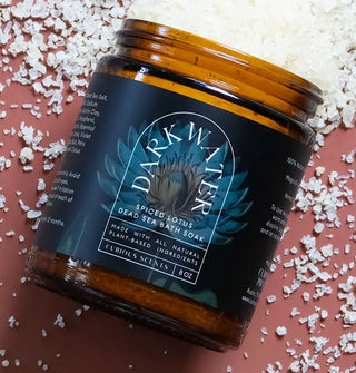 Amber jar of Dark Water Spiced Lotus Dead Sea Bath Soak with dark blue lotus and lily pad motif and white lettering rests on a terracotta-colored surface scattered with coarse salt