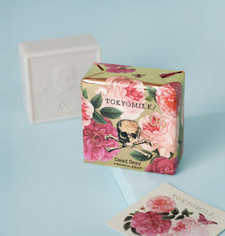 Square TokyoMilk Dead Sexy French Soap bar is wrapped in floral paper with a skull and crossbones drawing; a white unwrapped bar is behind it and a floral-patterned card is in the foreground