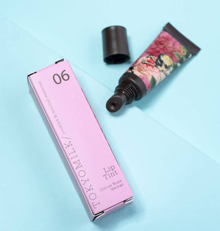 Opened tube of TokyoMilk Lip Tint in Citrus Rose Nectar scent featuring pink florals and skull and crossbones illustration next to its pink box packaging