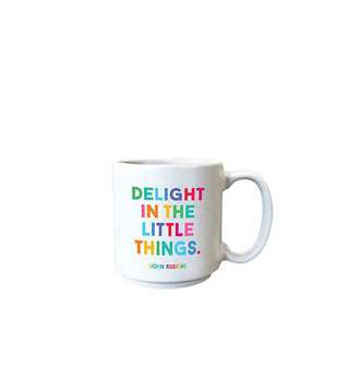 Small white coffee mug is printed with the John Ruskin quote, "Delight in the little things" in large multicolored lettering and the author's name below in smaller lettering