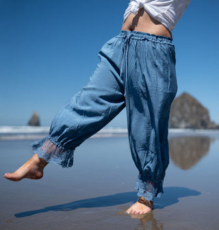 Model standing along the shoreline with sea stacks in the backround wears sky-blue, lace-trimmed pantaloons and kicks out one leg