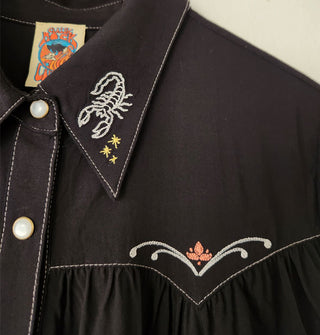 Closeup of scorpion, star, and flourish embroidery on the collar of a black button-down Western-style shirt with pearl snaps