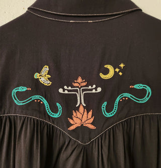 Closeup of colorful snake, hawk, moon and star, and floral embroidery on the back yoke of a black Western-style shirt with gathering below the contrast-stitched seam