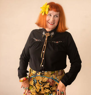 Smiling model with a red bob wears a black Western-style shirt with detail embroidery, intricately patterned pants, turquoise rings, a bolo tie, and a yellow flower in hair