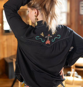 Model facing away from the camera lifts hair to reveal colorful embroidery on the back yoke of a black Western-style shirt