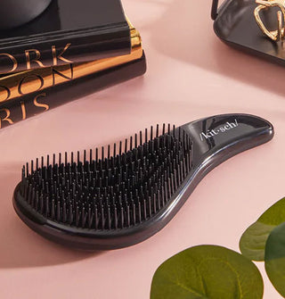 Curvy black hairbrush with "Kitsch" printed in white on the handle rests on a pink surface with a stack of black and gold books and other items