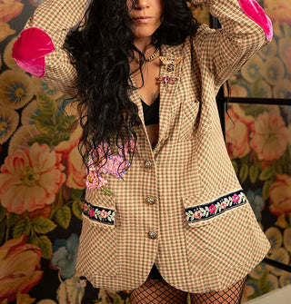 Model standing in front of a lush floral wallpapered backdrop with mirror wears a beige and green micro-plaid oversized blazer with bodice appliqué and pocket trim, brooches attached to the left lapel, and pink velvet elbow patches, paired with black fishnet stockings